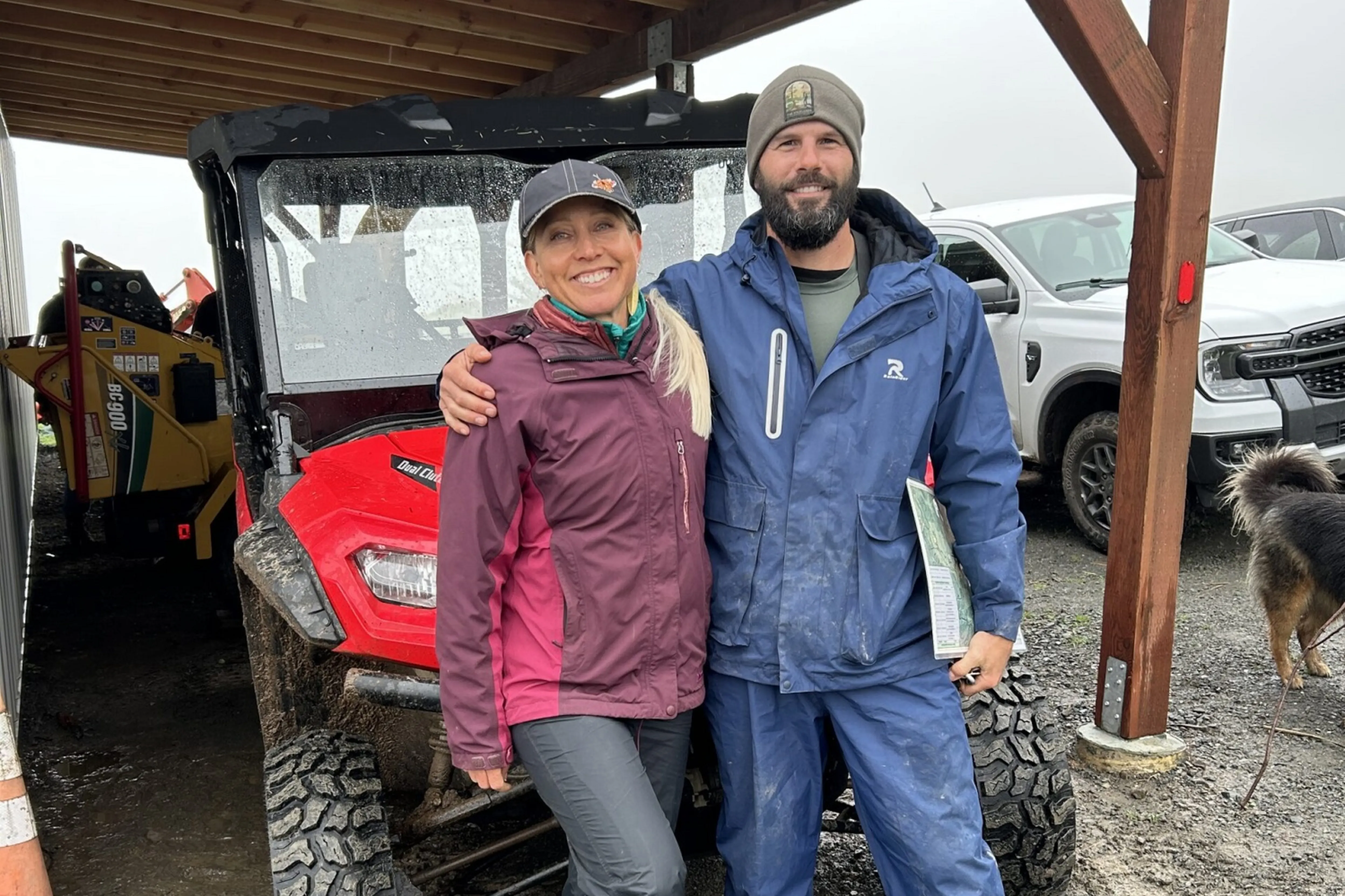 A man and woman in outdoor gear stand smiling under a wooden shelter beside a red ATV. A dog and parked vehicles are visible in the background.
