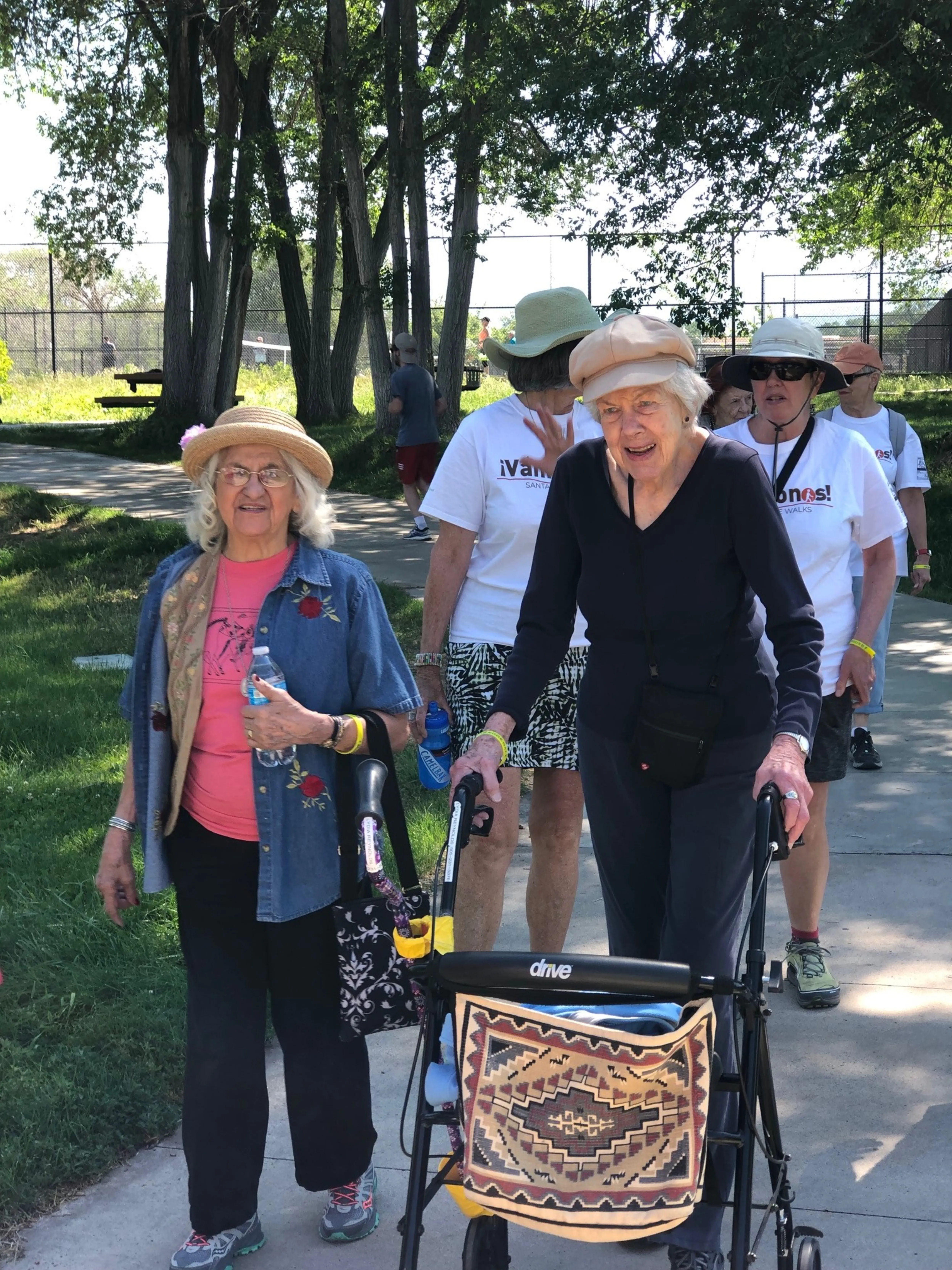 Older women outside walking on a sunny day. One woman leans on a walker with wheels to assist her.