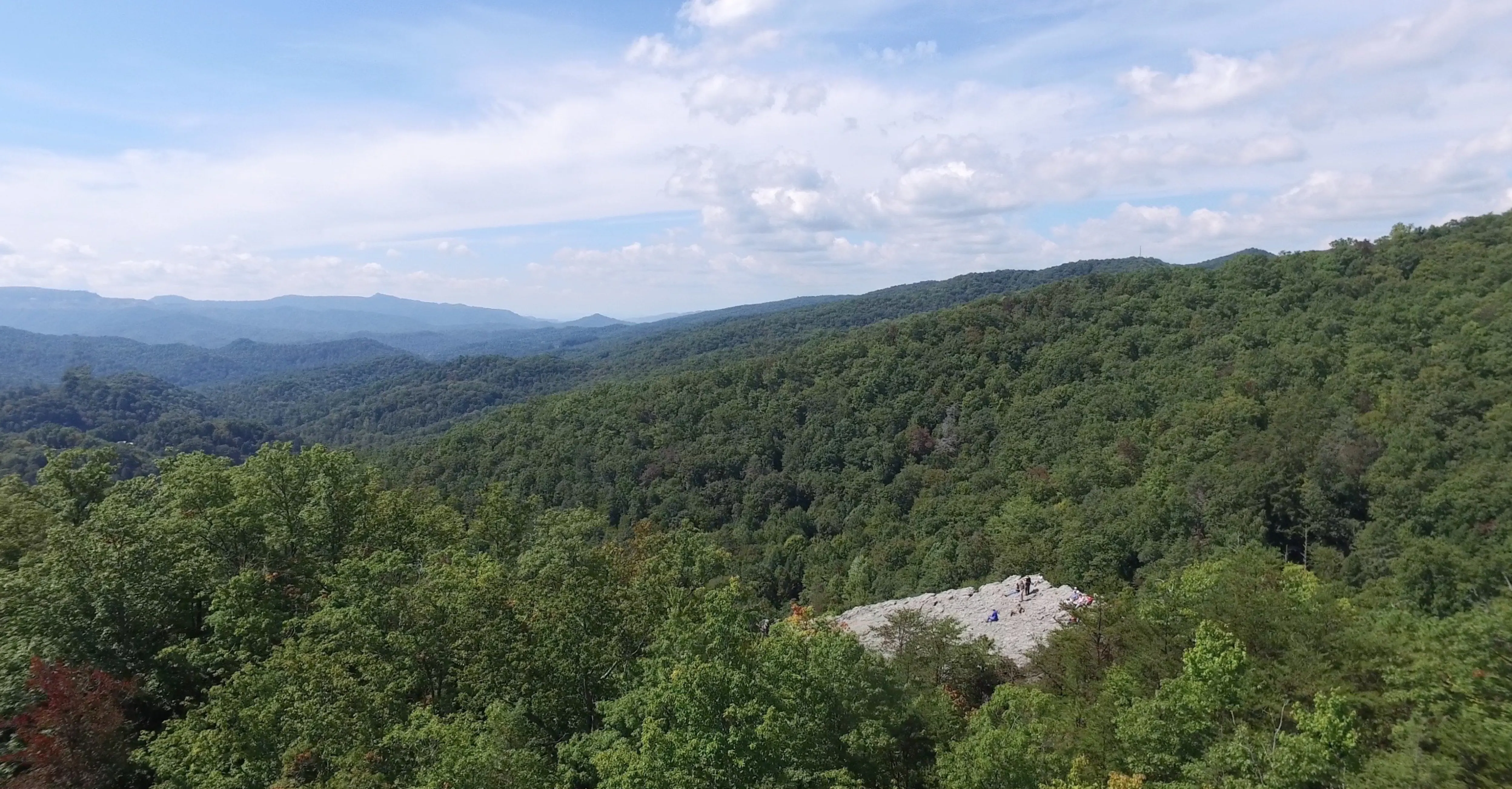 Overhead view of mountain range covered in green trees, with a small patch of rocks where it looks like people are sitting