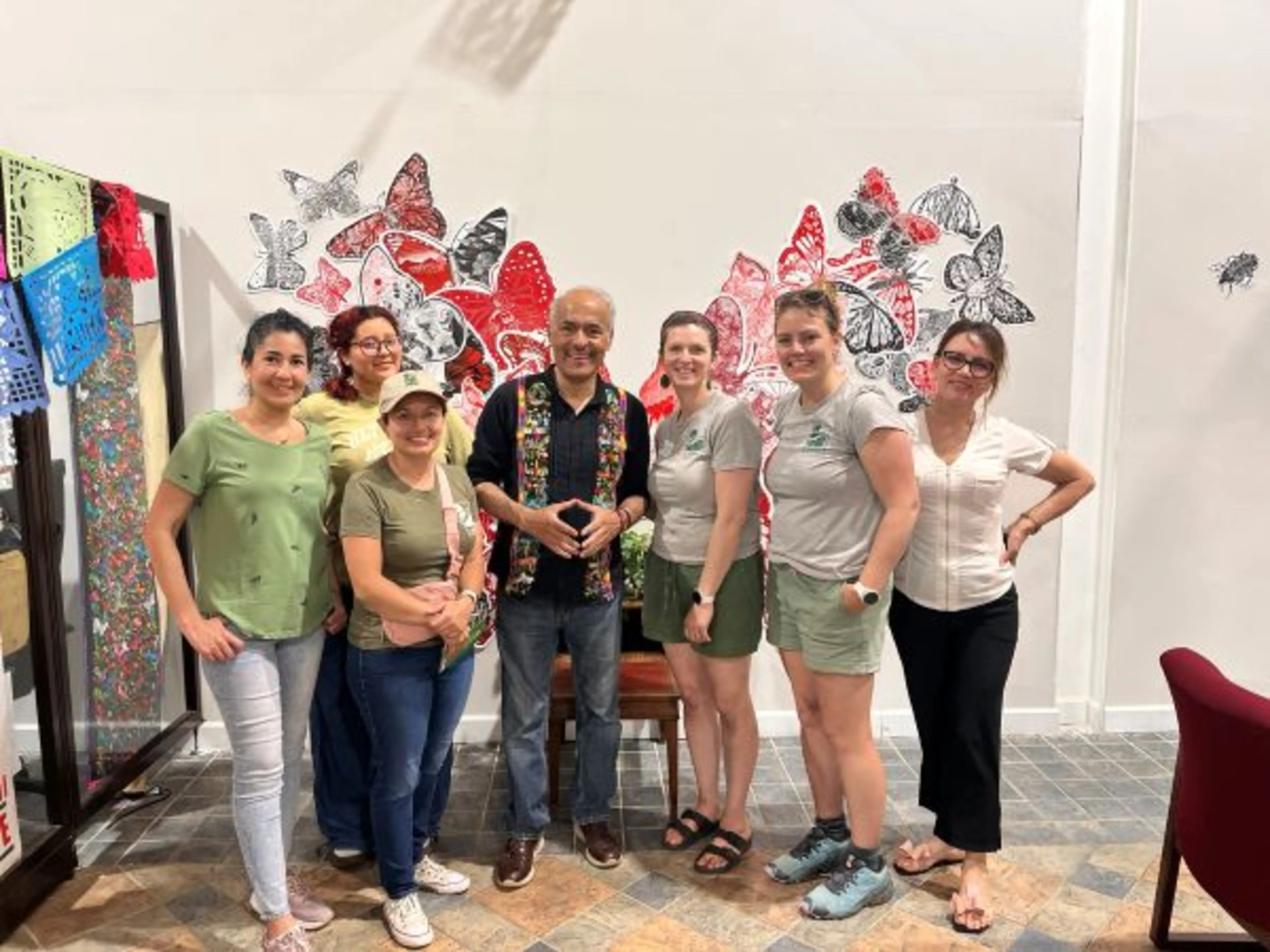 A group of seven people smiling indoors, standing in front of a butterfly-themed mural. Some wear casual green shirts and one is seated.