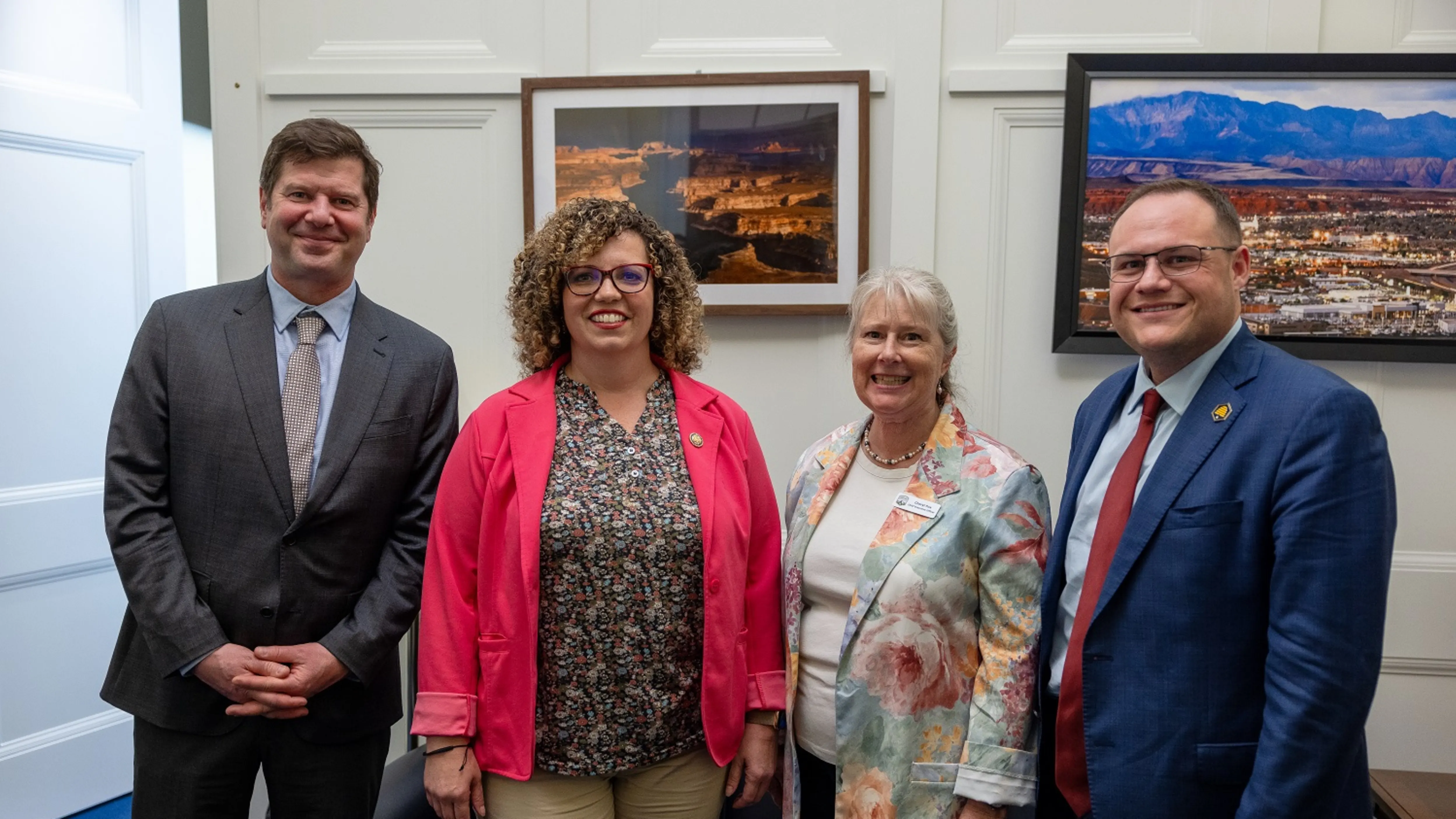 Four people standing in a room with framed landscape photos, dressed in business and smart casual attire, smiling at the camera.