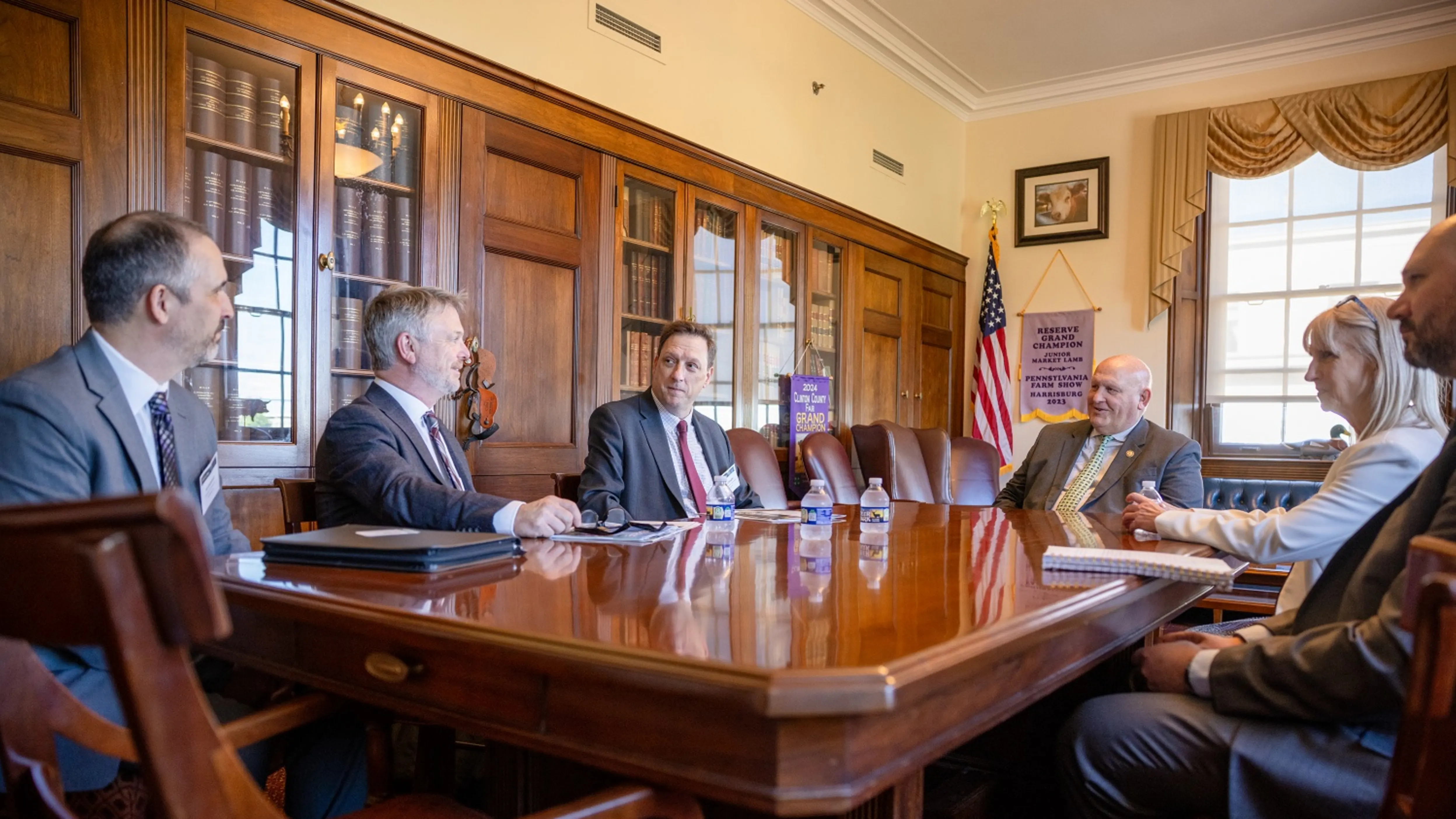 Six people in formal attire having a meeting around a wooden table in an office with bookshelves and an American flag.