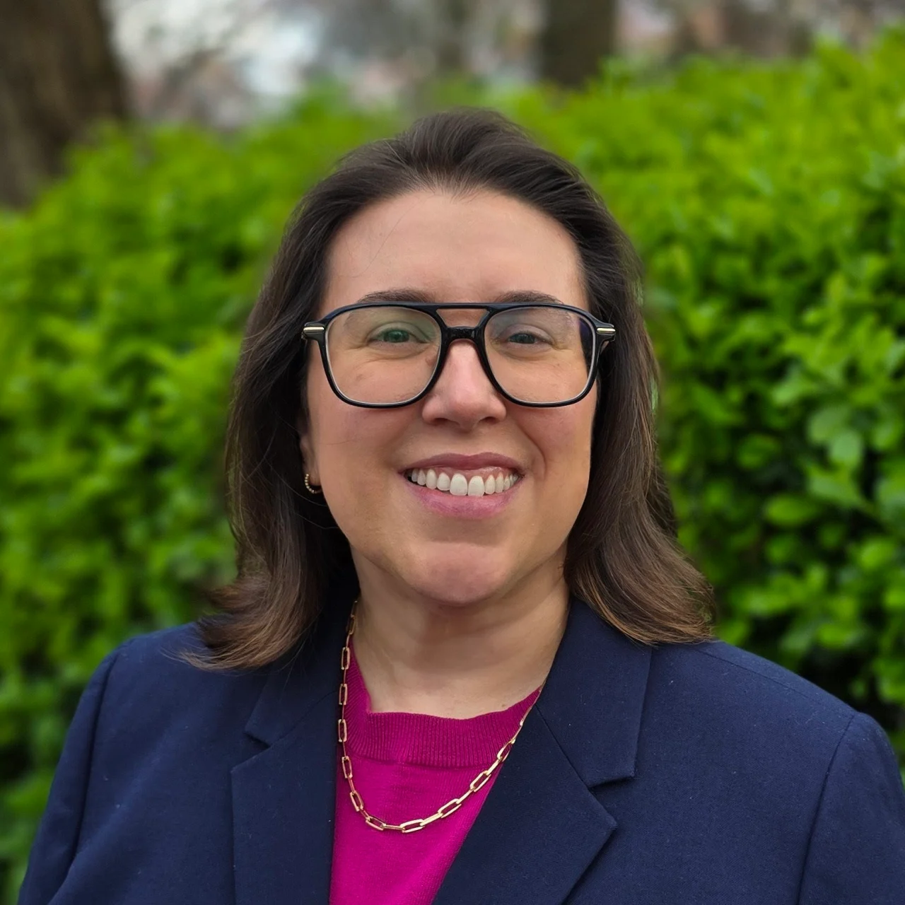 Woman wearing glasses and blazer smiling in front of bushes