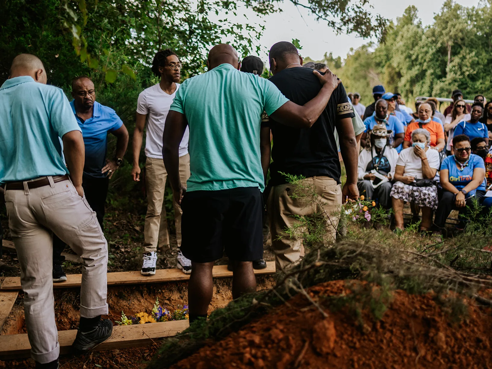 Six men standing around a grave looking down into the hole, standing next to a pile of dirt, with people seated in the background, looking as if they're crying.