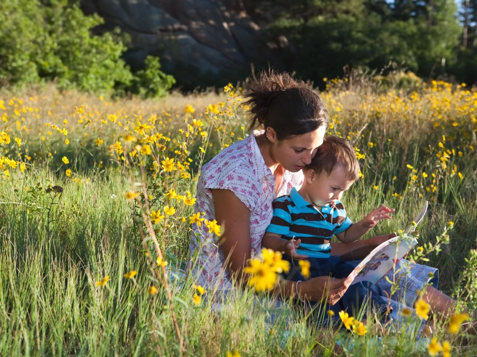 A woman sits in a field of flowers with a child in her lap reading a book