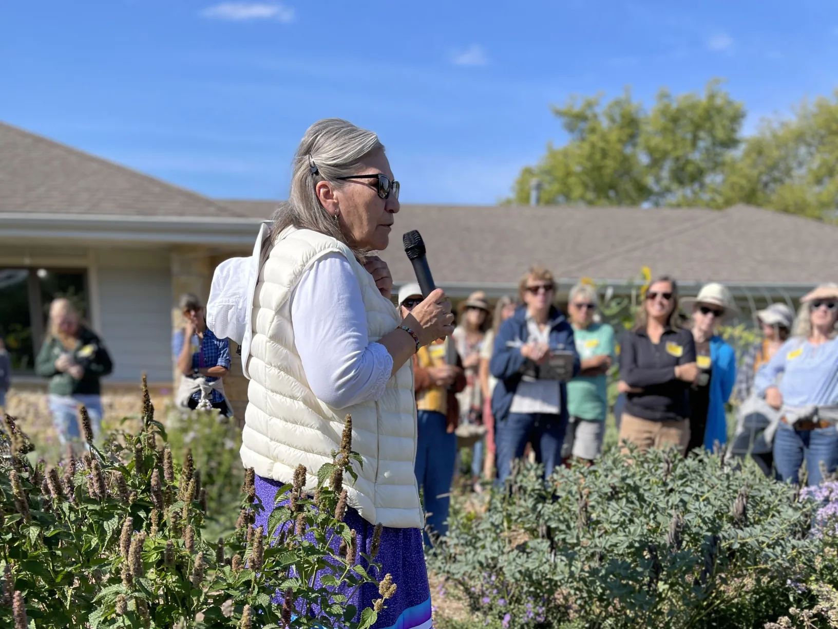 Woman standing in a garden with a microphone in front of a group of women.