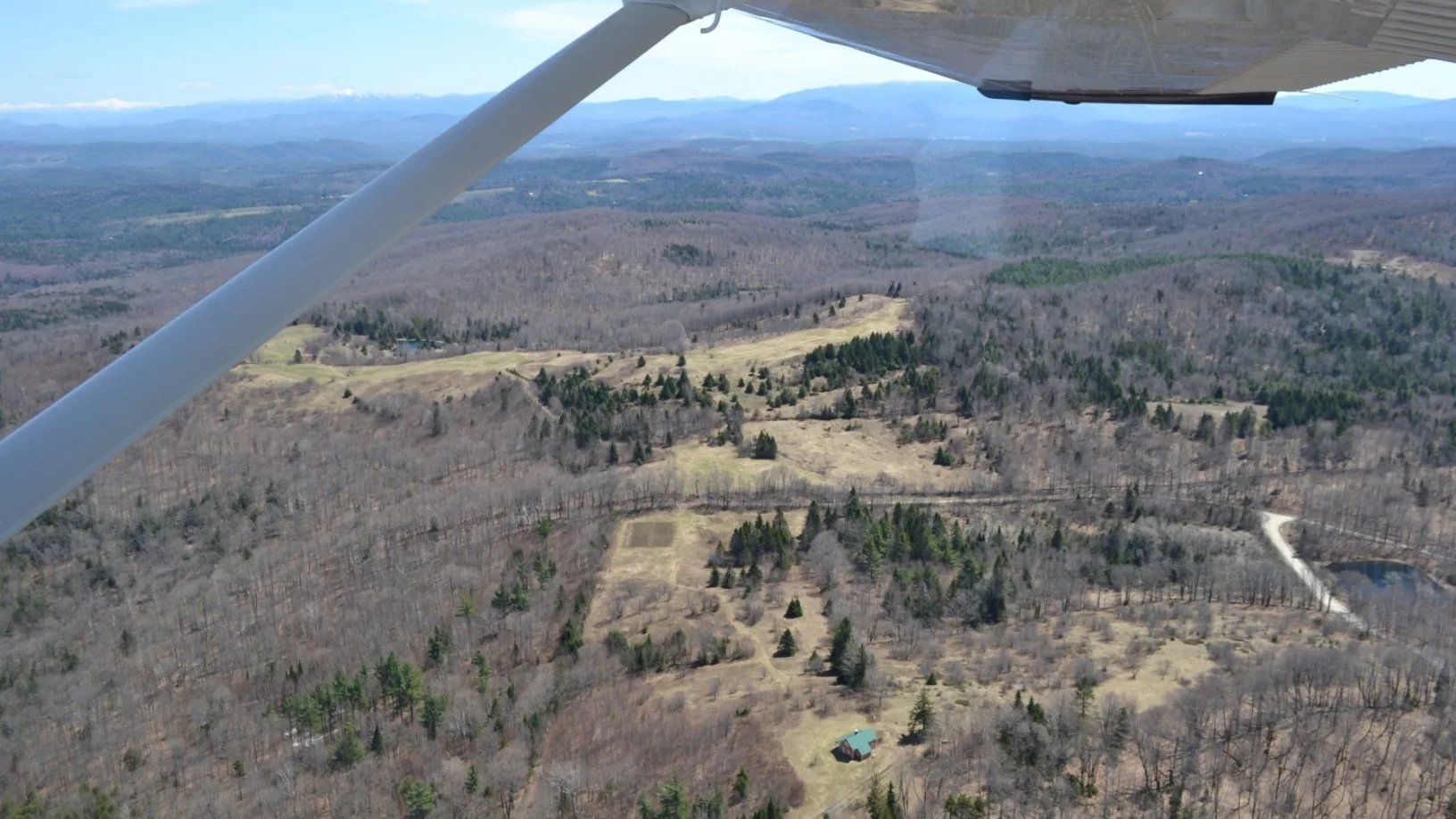 Looking out a plane window down at brown fields with some green trees scattered and a house on it.