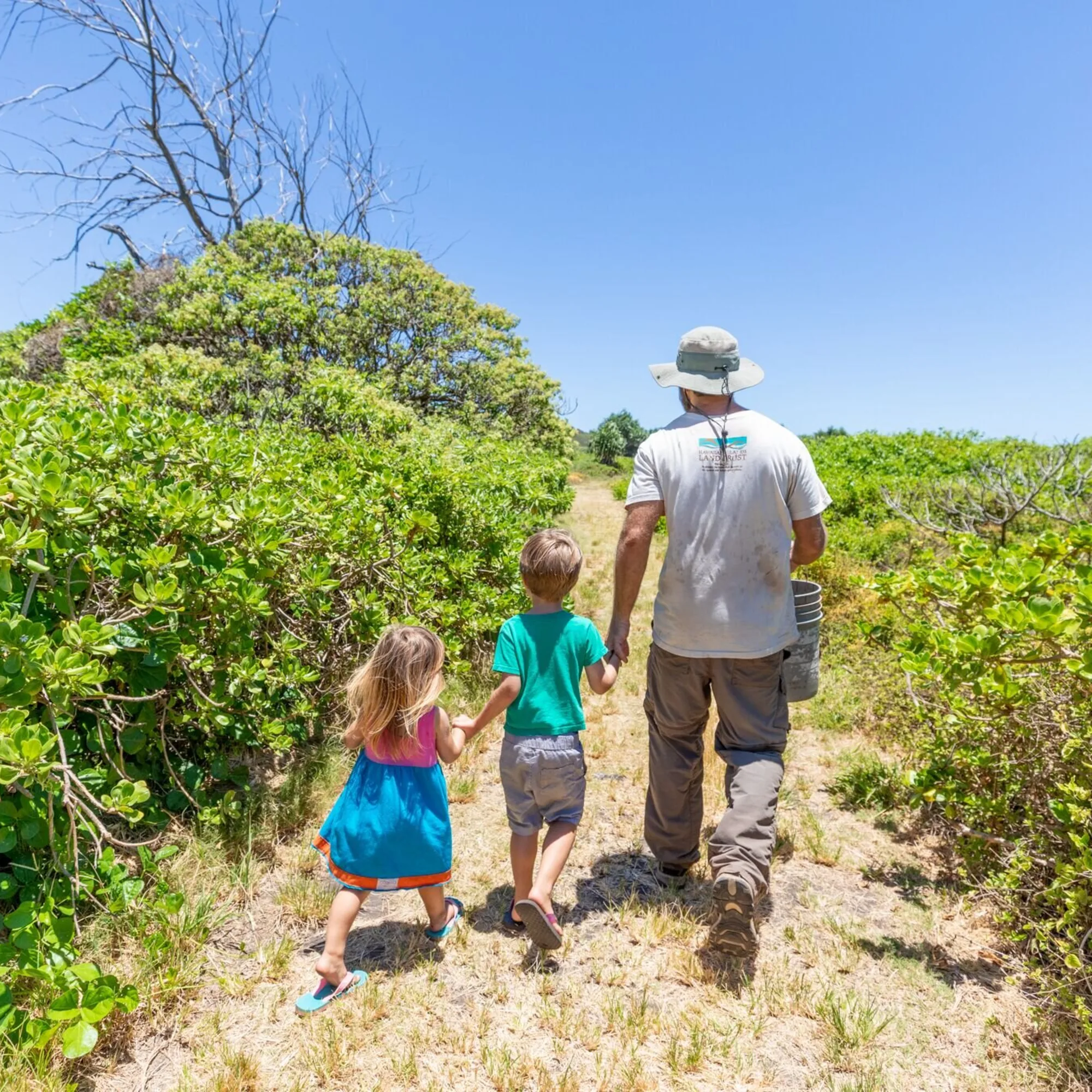 A man holding a bucket with two children walking down a path surrounded by trees.