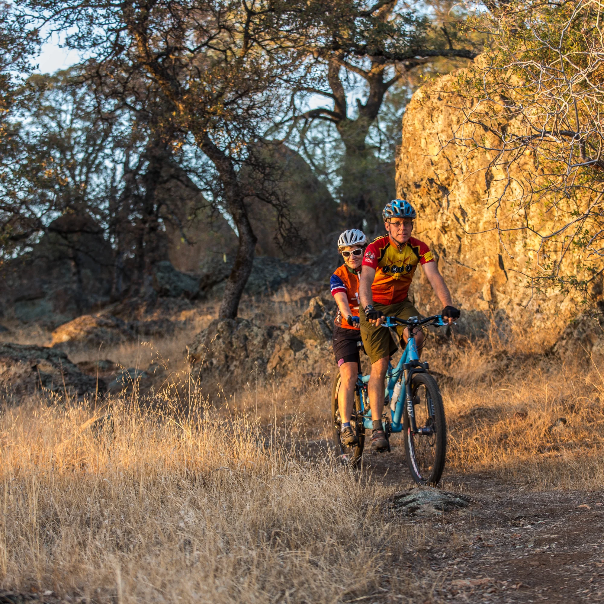 Two people riding on a tandem bike on a trail surrounded by trees and large rock formations