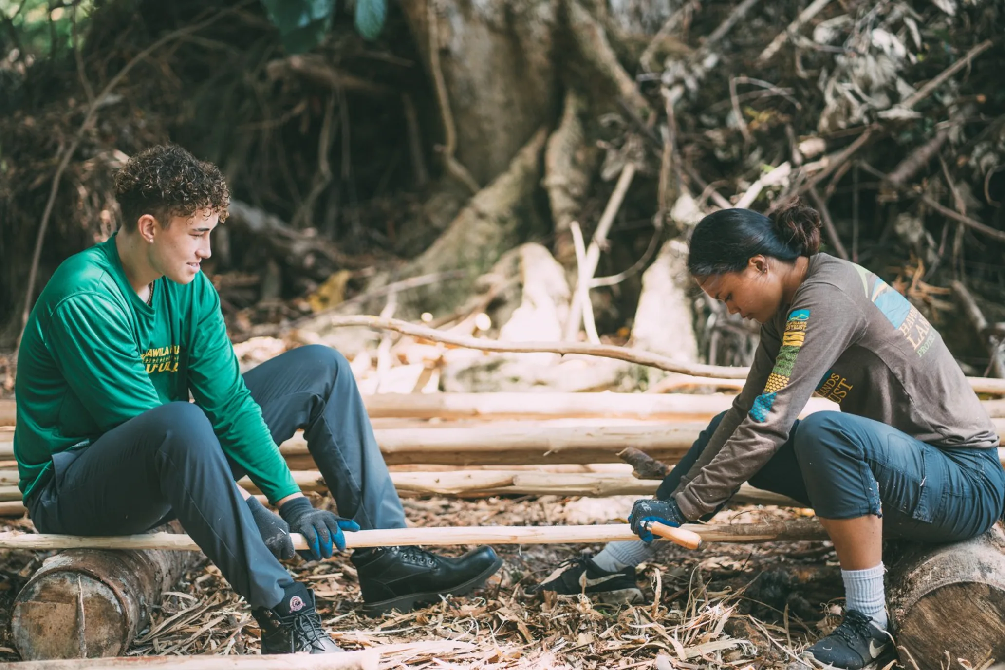 Two people sitting holding a piece of wood between them, one holding a tool on the wooden stick