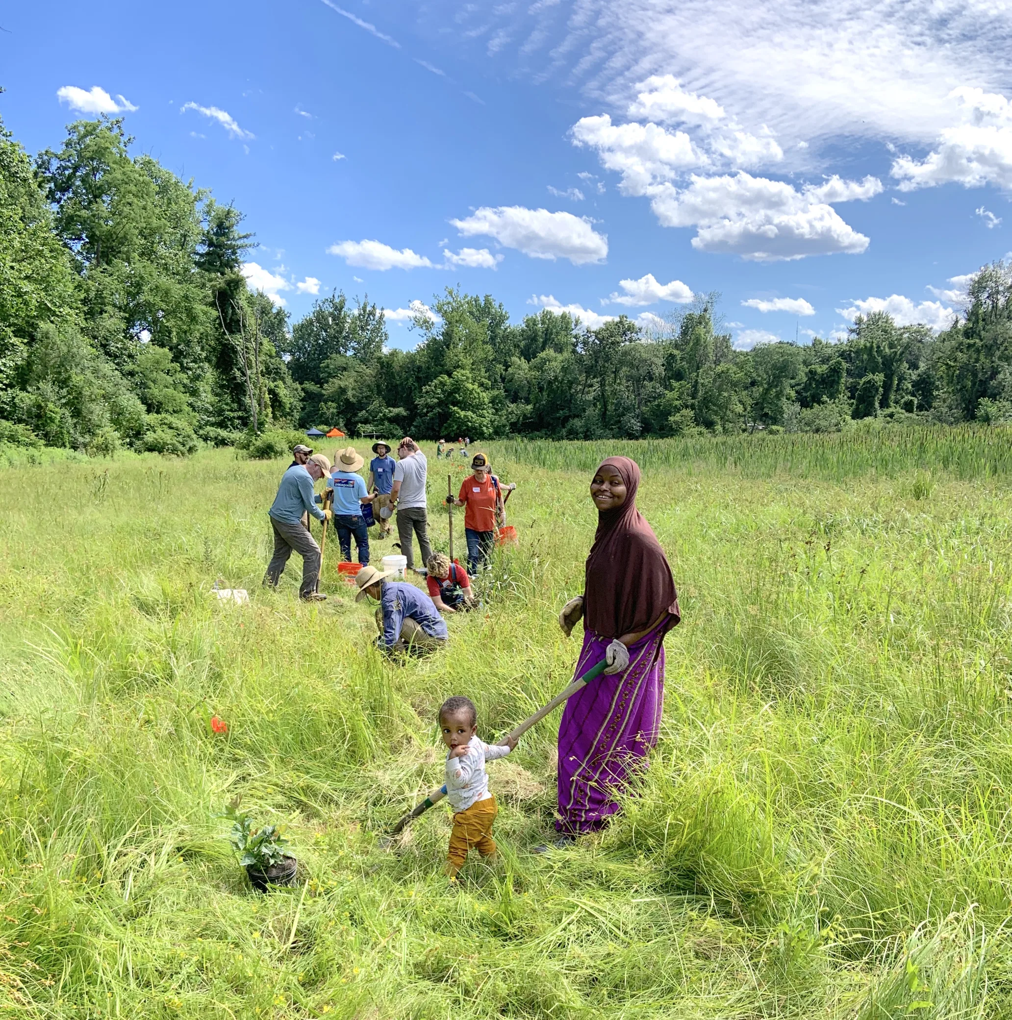 A woman wearing a hijab holds a gardening tool with a small child next to her, both looking at the camera. Behind them are other people with various tools working in a field.