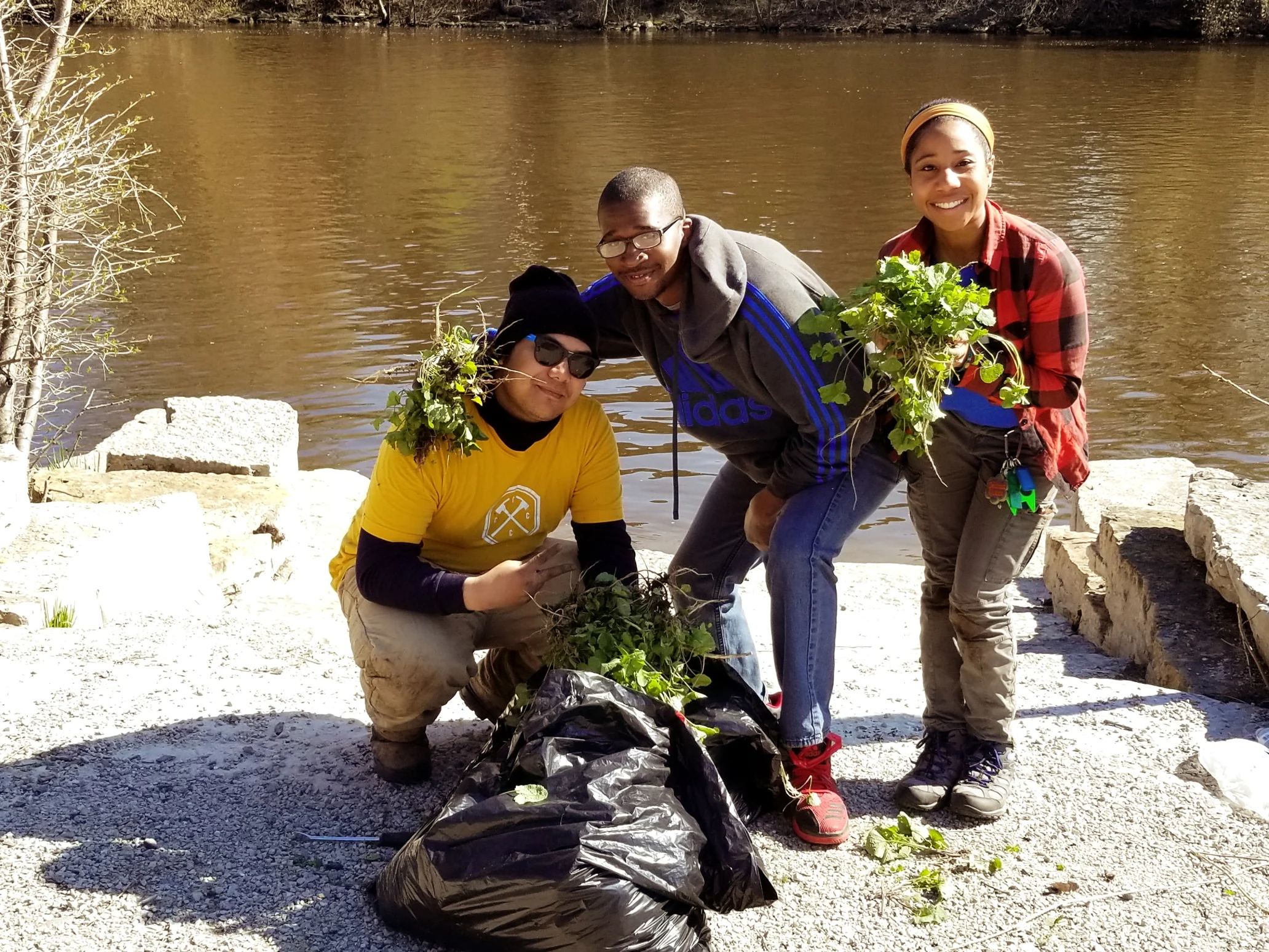 Three people with a trash bag and holding leaves standing in front of a river.