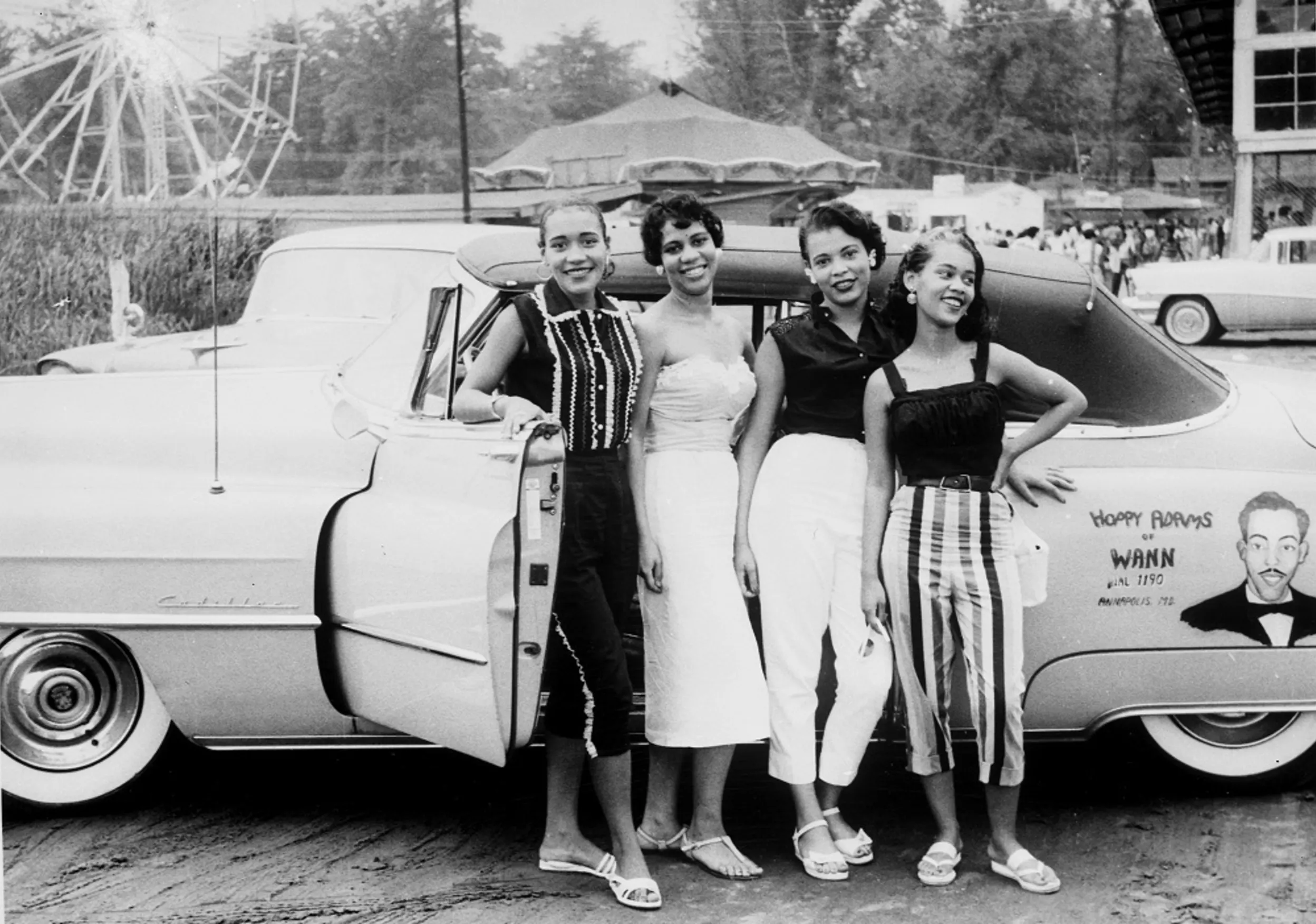 A black and white photo of four young Black women standing outside their car at the edge of Elktonia Beach, which welcomed Black people in the 1930s through 1970s.