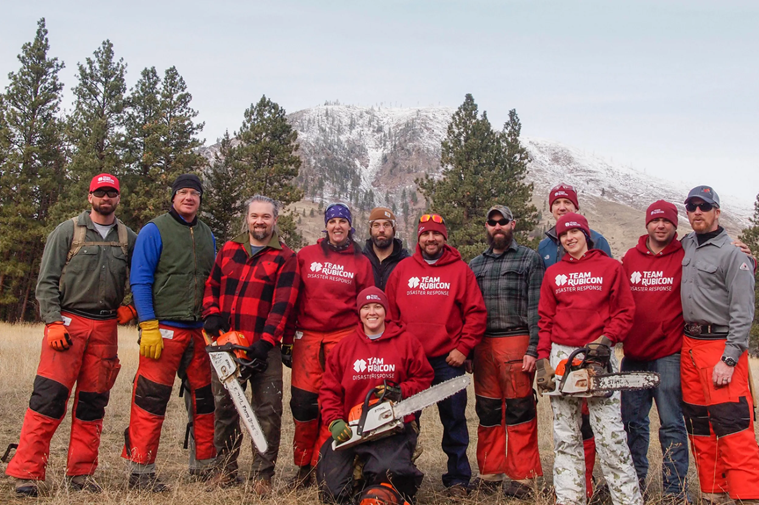 Military veterans, many in red Team Rubicon sweatshirts, stand for a group photo with their chainsaws, with mountains and pines behind them.