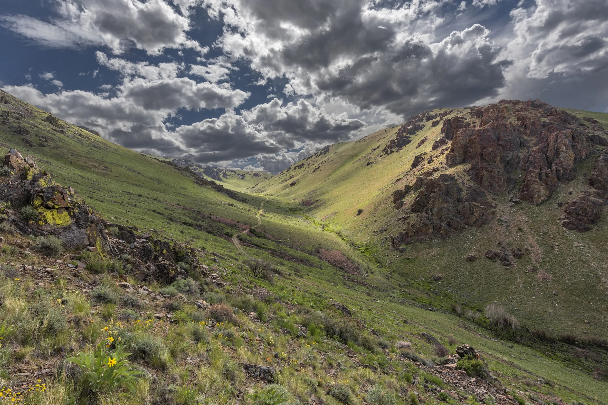 Grassy mountains give way to rocky mountain tops, split by a shallow valley between as the sun shines through thick white clouds above.