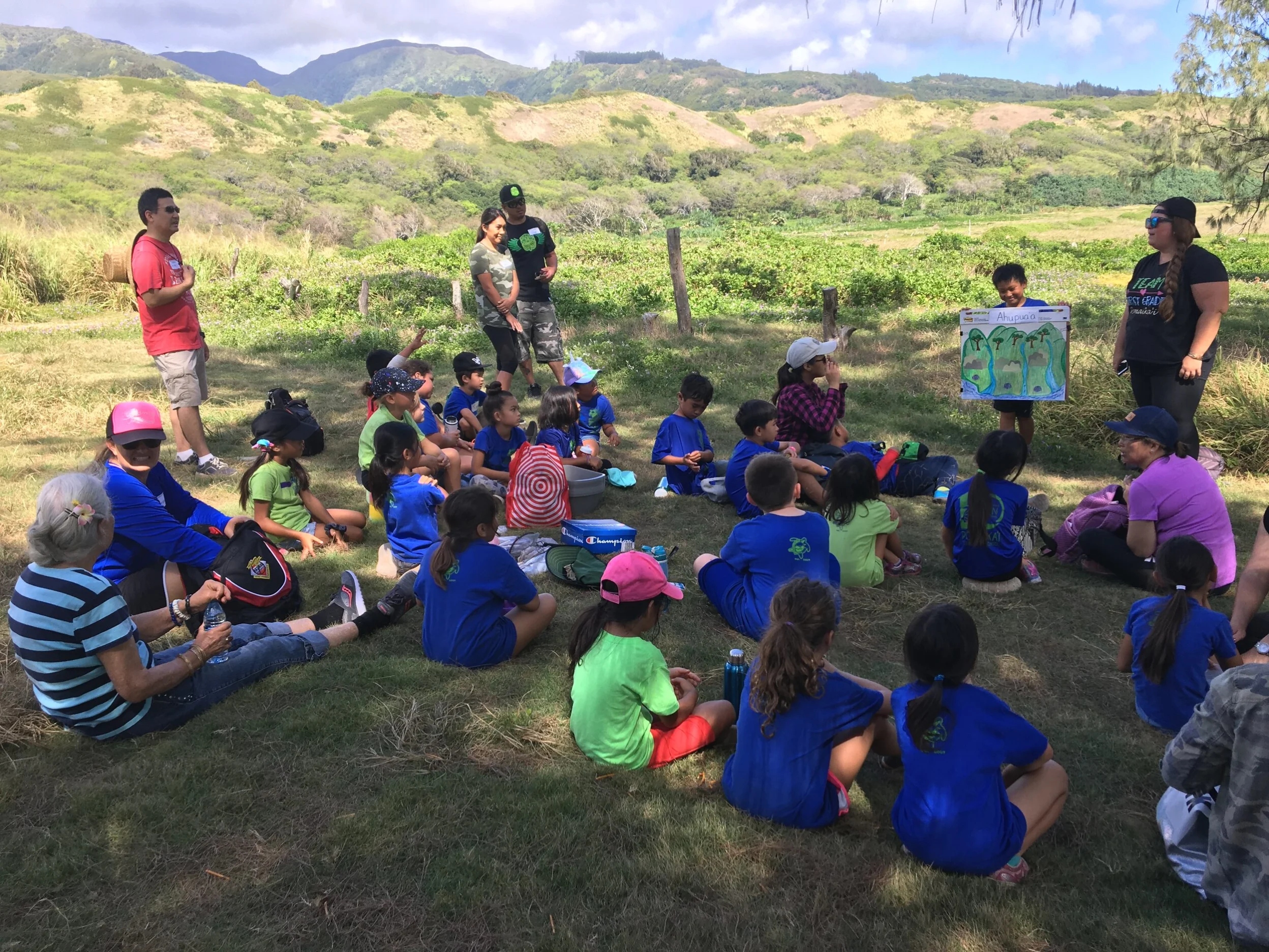 Children sitting on the ground with some adults, with a child standing at the front of them holding a large poster board. They are in a field surrounded by green hills.