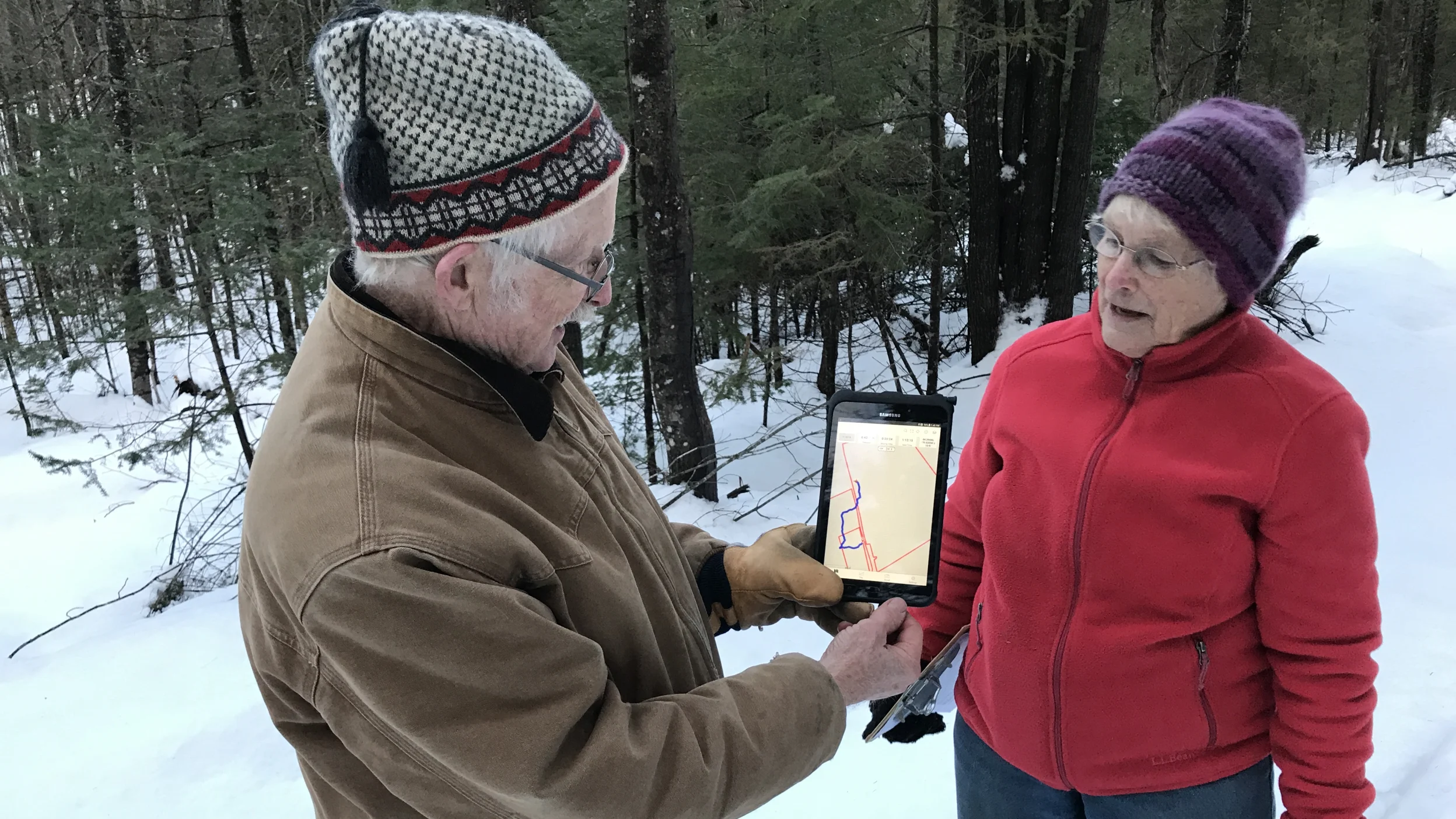 Two people standing in a snowy forest looking at a tablet