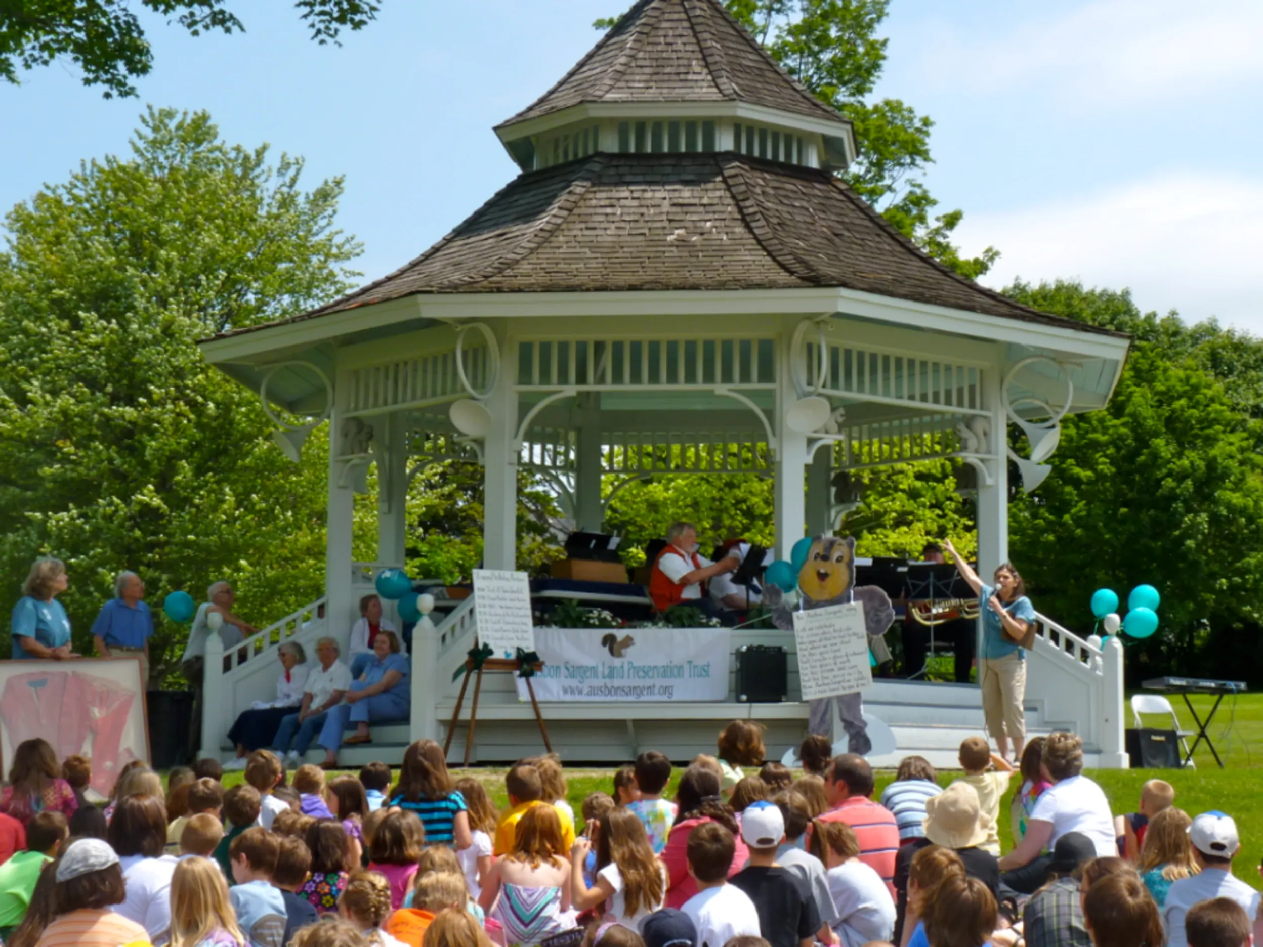 A gazebo with a band inside of it playing with children and families sitting around it.