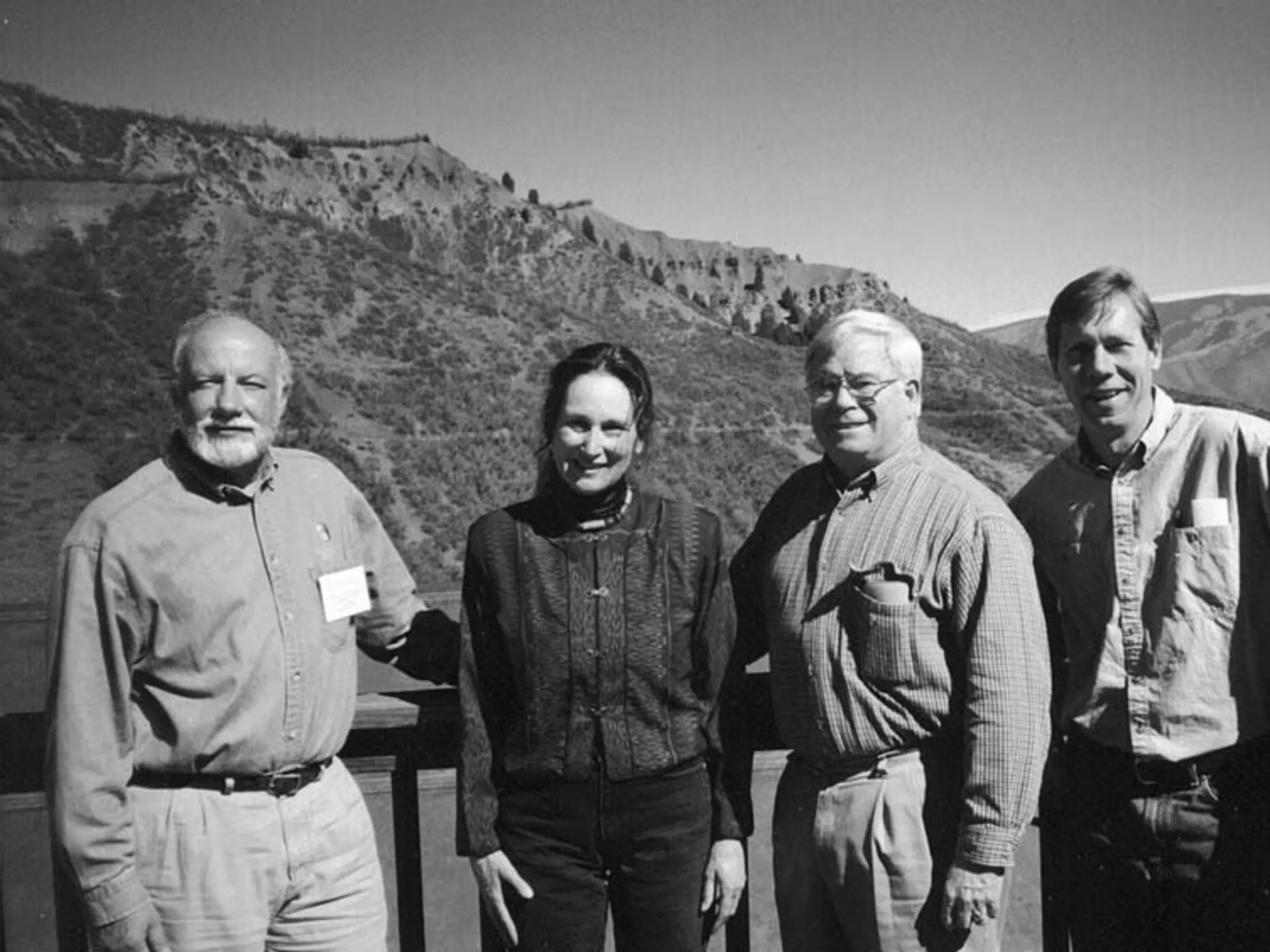 A black and white photo of three men and one woman standing on a deck with a mountain in the background.