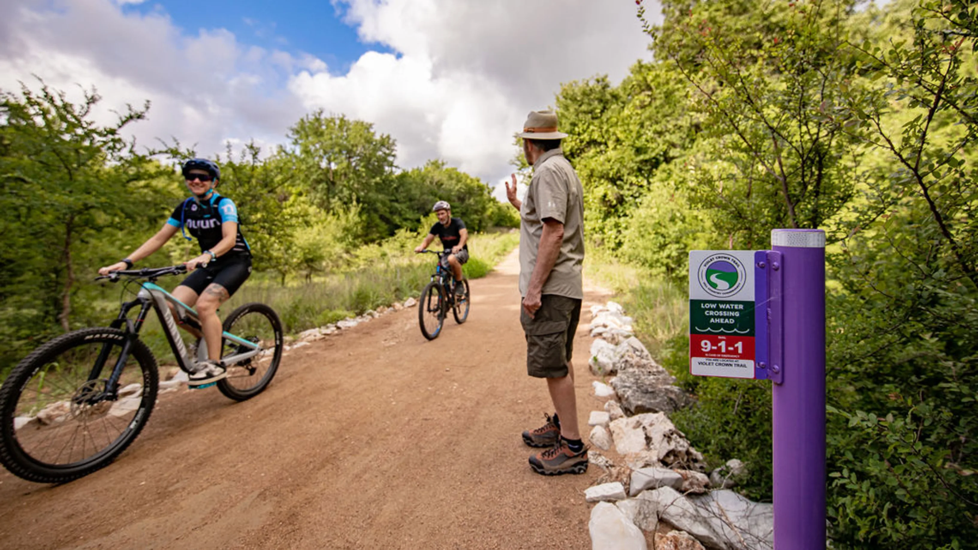 Two bikers biking down a trail surrounded by trees, with a person in a hat and khaki outfit waves at them.