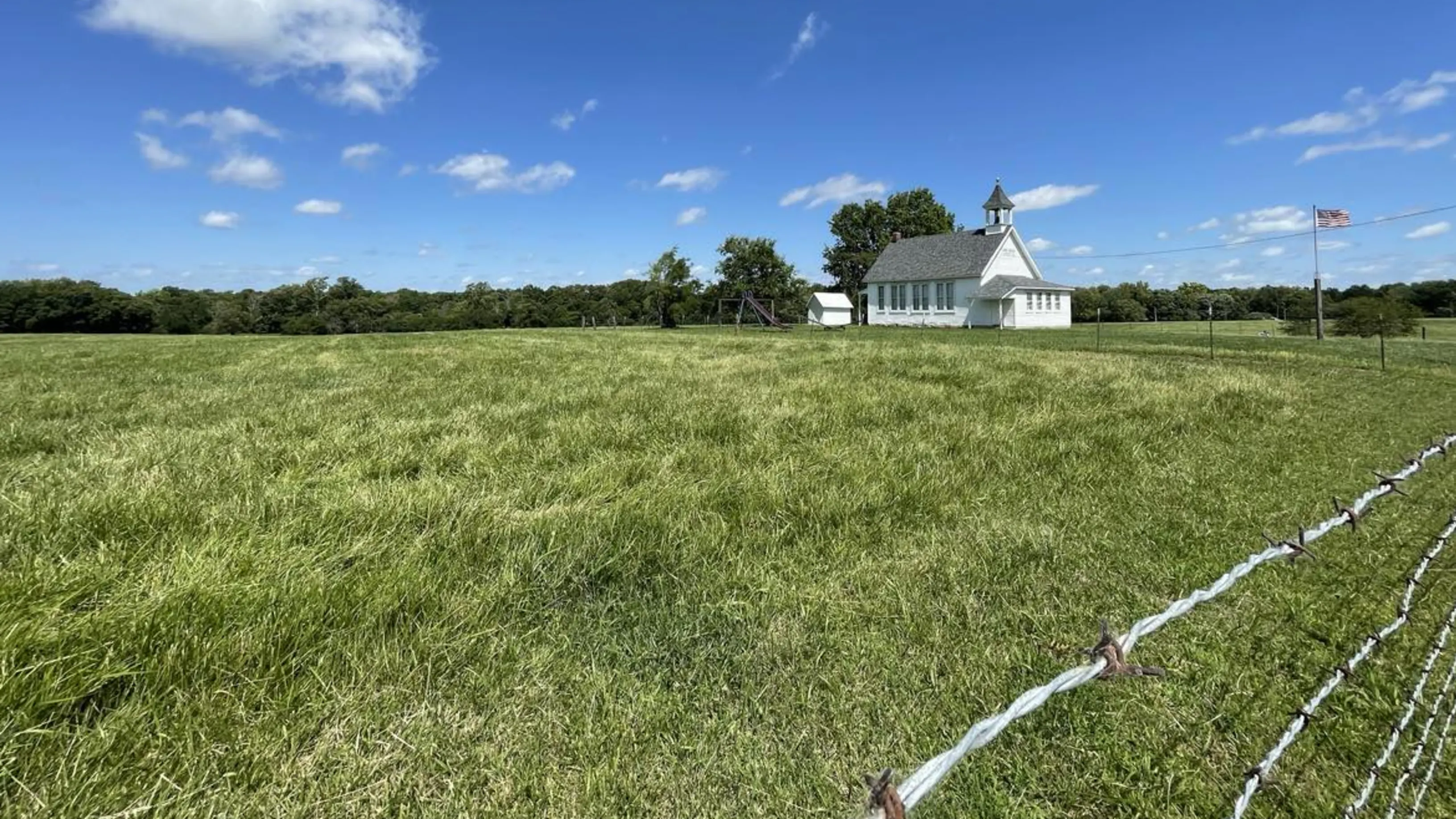 A white schoolhouse with a flag in front surrounded by green grass.
