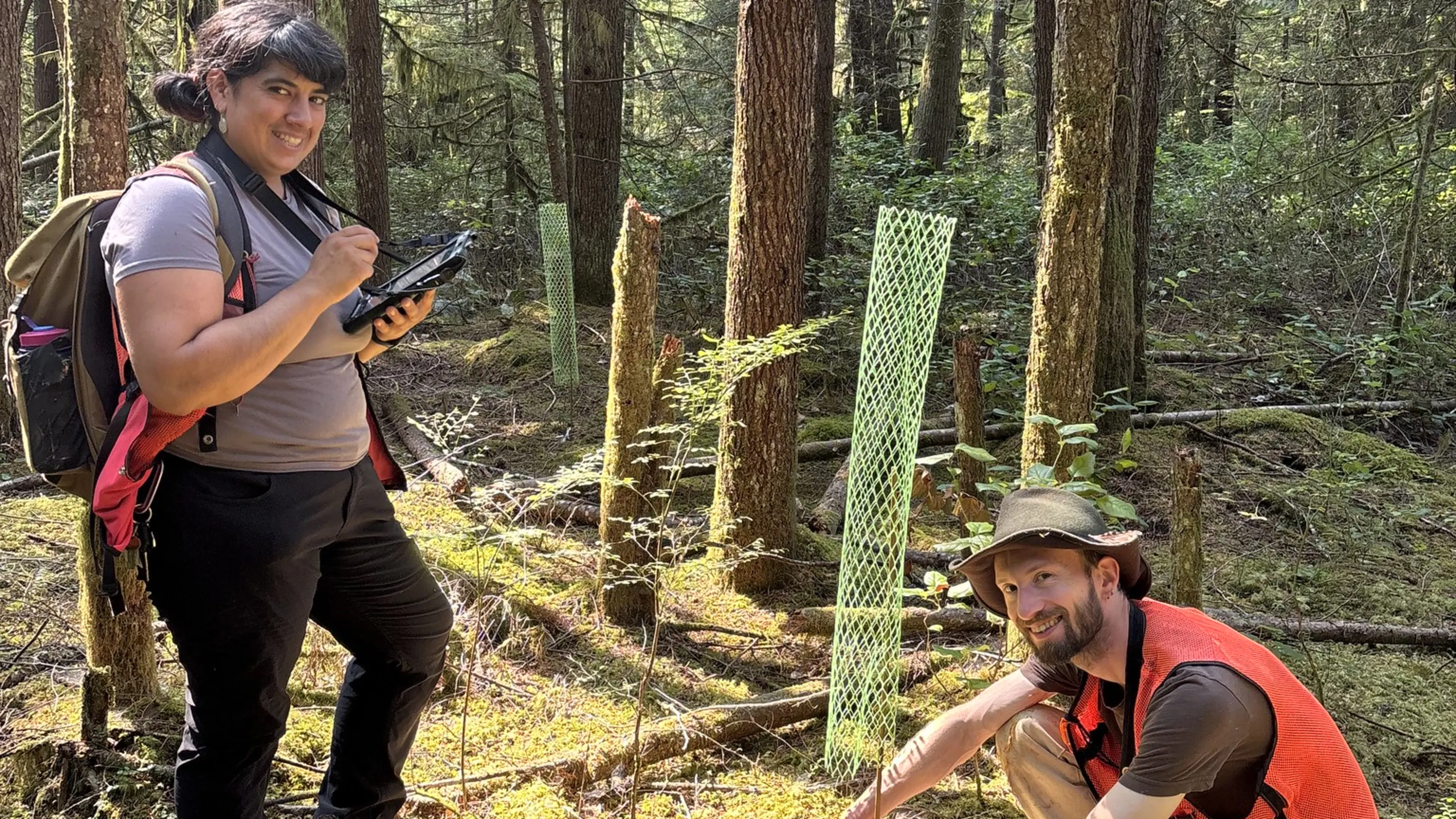 One person wearing an orange vest kneeling in front of a sapling with mesh around it. A second person stands with a iPad next to the sapling, both smiling at the camera. There are trees behind them.