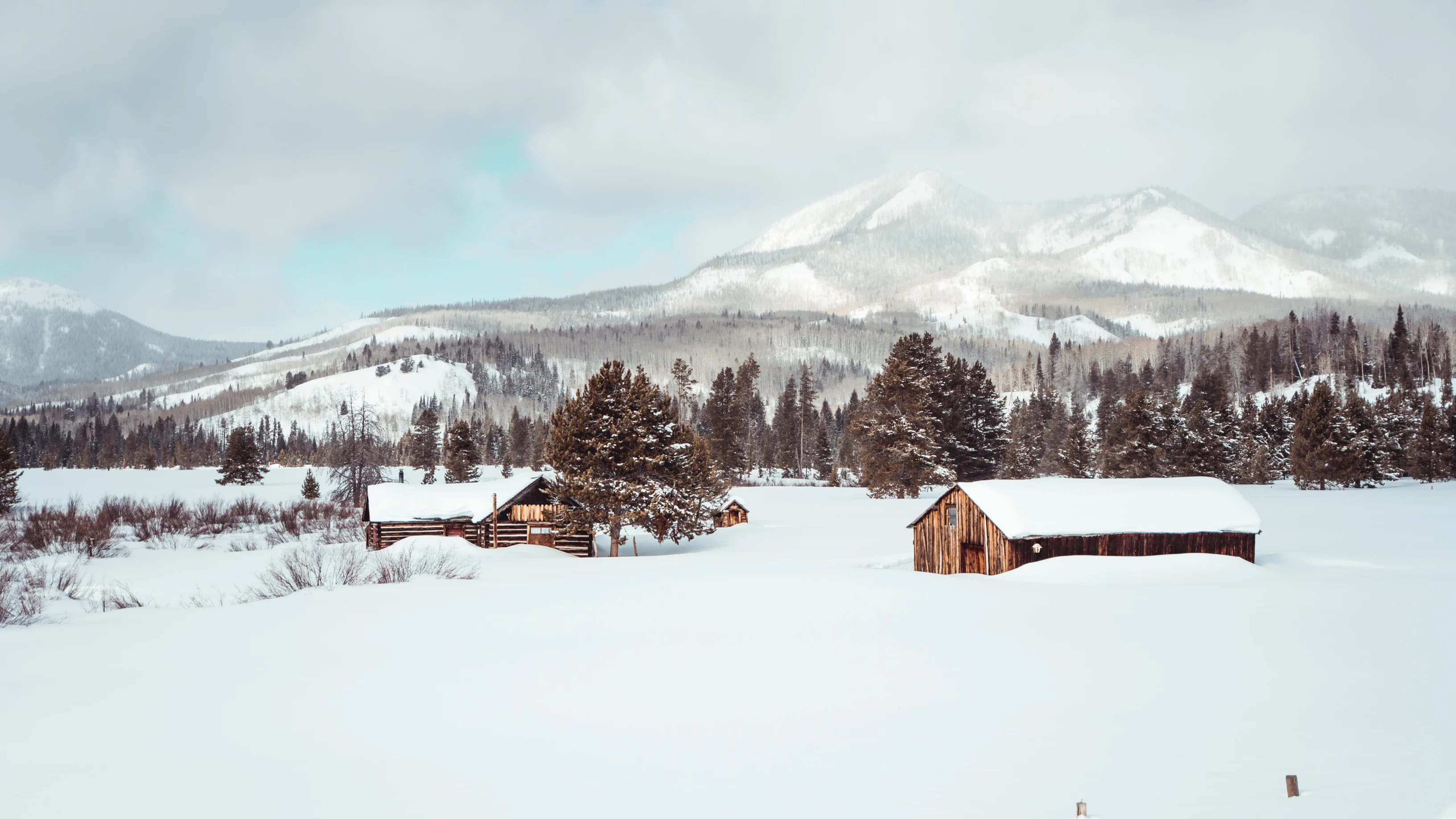 A ranch covered in snow with two building on the property, surrounded by trees. A mountain in the background.