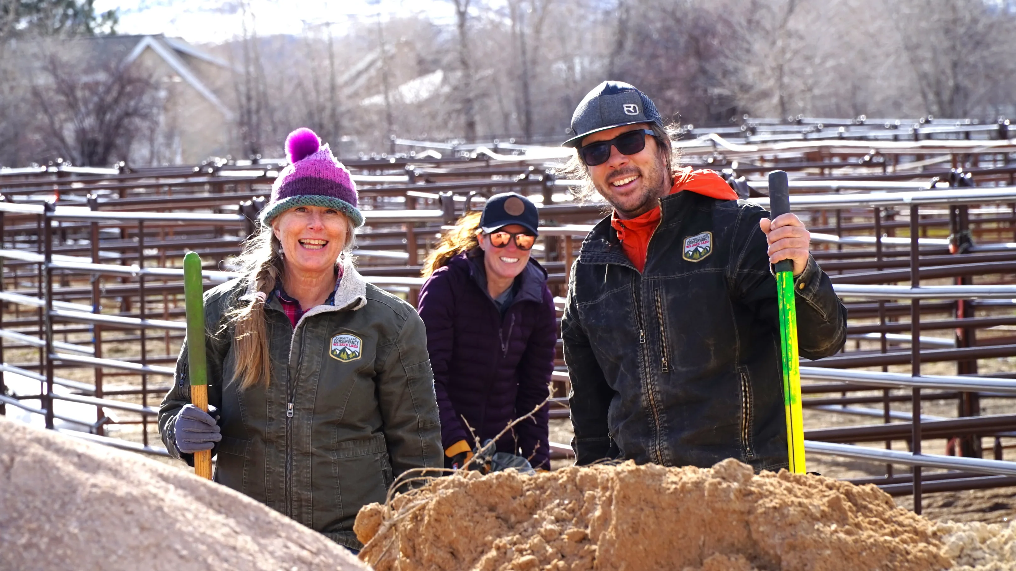 Three people wearing work clothes holding shovels in front of cattle pens.