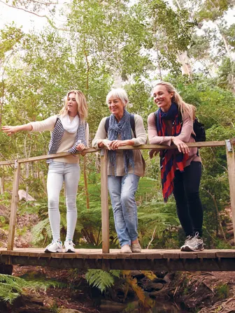 Three people stand on a bridge in a forest, two who are younger in age and one who looks older.