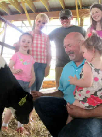 Man kneeling holding a baby while petting a baby cow, with other children and adults surrounding them smiling