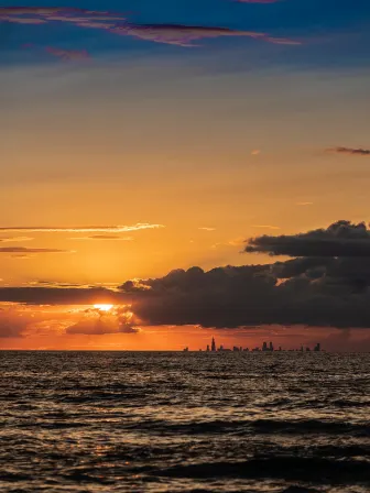 Photo of the sun peeking out behind clouds, over a large body of water, with a city scape next to it.