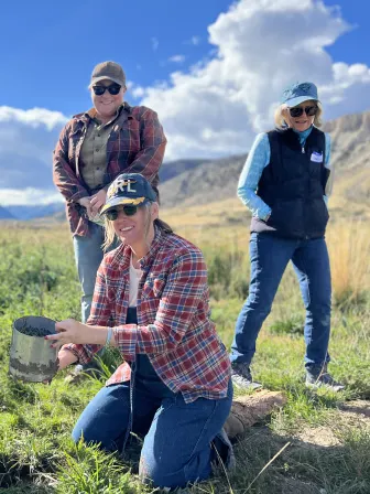 Four women in a field wearing hats and working gear, one holding a bucket and one with a shovel.