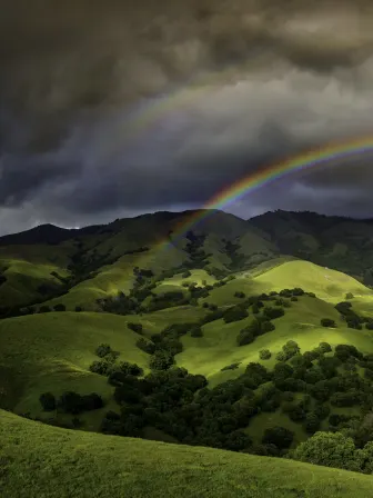 Rainbow over dark clouds, above rolling green hills with trees covering it.