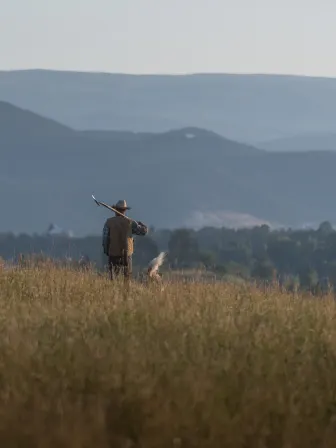 A man standing in a field with his back to us, holding a shovel over his shoulder looking out towards a distance mountain range.