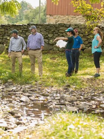 five people stand in a circle in front of a creek while one holding a clipboard points at something