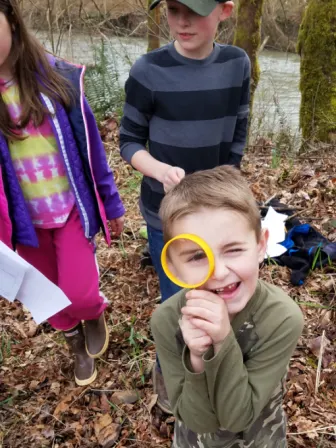 Child holding a magnifying glass against eye, with two other children behind them.