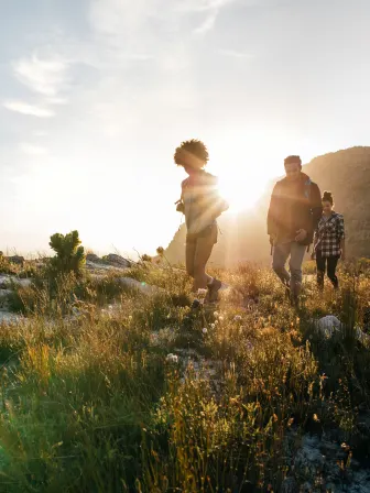 Three people walking on a grassy mountain with the sun low in the sky behind them.