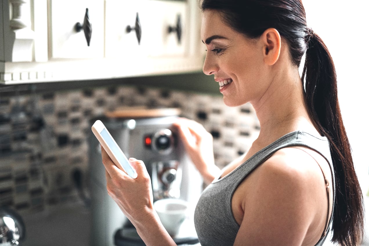 Smiling woman is reading from a phone, standing near her espresso machine