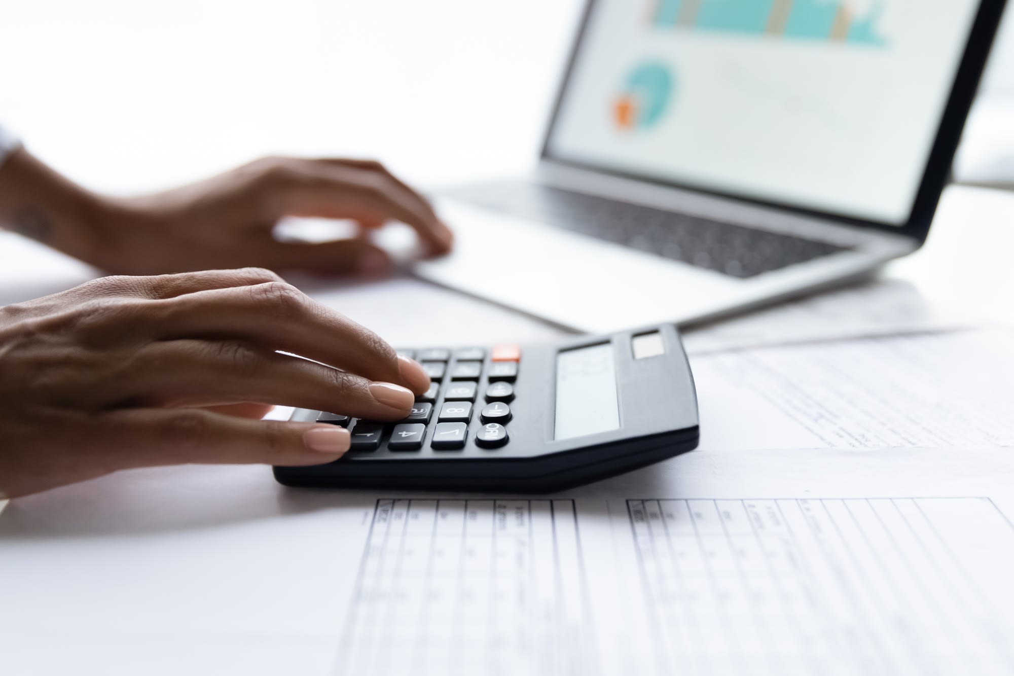 Individual multitasking with a calculator and laptop at a desk covered in financial documents