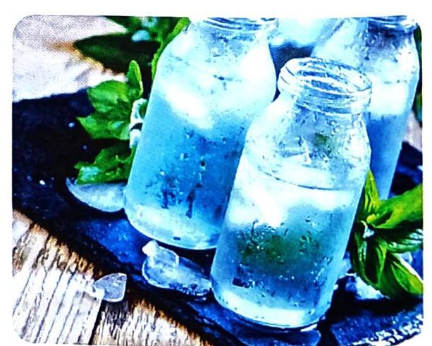 Three condensation-covered glass bottles filled with blue liquid and ice, with fresh mint leaves scattered on a dark, textured surface.