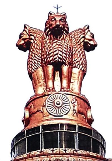 The State Emblem of India features four lions, with three heads visible in a triumphant posture, atop an abacus. The abacus is adorned with the Ashoka Chakra in the center, flanked by a galloping horse and a bull on its sides, with another lion visible. The sculpture is rendered in a metallic, bronzed finish.