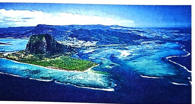An aerial view showcases a steep, dark volcanic peak covered in vibrant green vegetation, surrounded by clear turquoise waters. The shallow water reveals intricate patterns of coral reefs. Distant, hazy blue mountains form the backdrop under a bright blue sky with scattered white clouds.