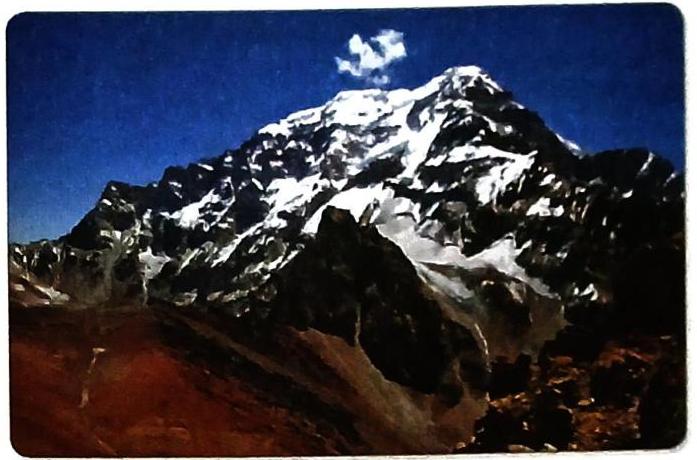 A snow-covered mountain peak under a dark blue sky, with a single fluffy cloud near the summit. The mountain's lower slopes are rugged and reddish-brown, while the upper reaches are dominated by snow and ice, revealing jagged rock formations.