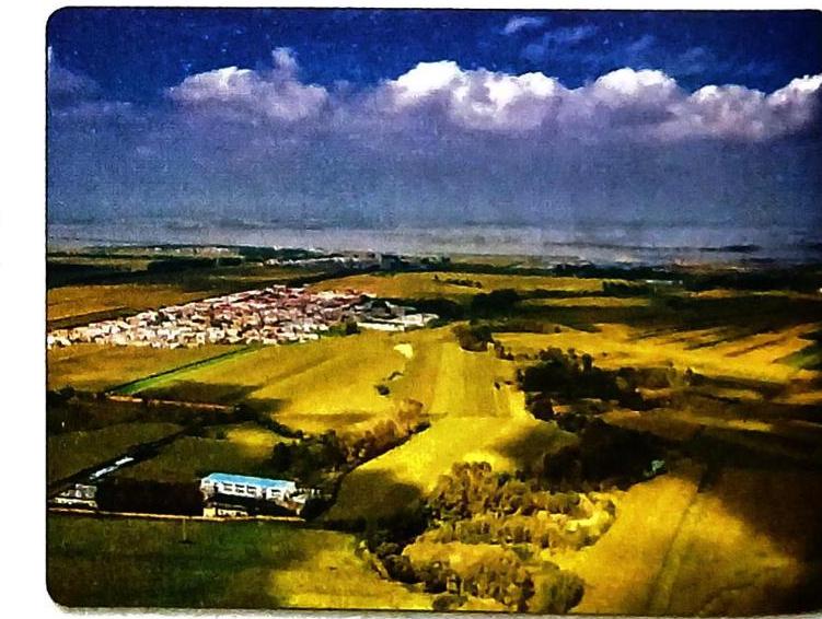 An aerial view captures a landscape under a blue sky with white clouds. A settlement of numerous white buildings is situated on rolling fields of green and golden hues, marked by shadows. In the foreground, a long, rectangular building with a blue section stands amidst fields. Scattered trees dot the scene, contributing to the rural panorama.