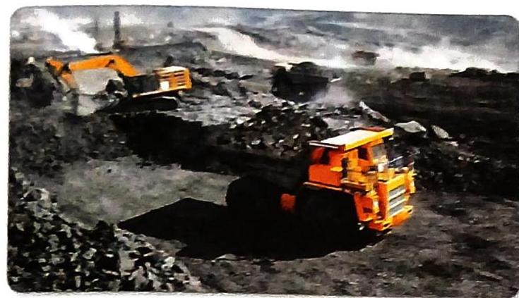 An orange excavator loads material into a large orange dump truck at a dark, excavated site. Piles of dark material are visible, and other heavy machinery operates in the background amidst dust or smoke.