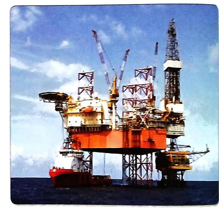 A large offshore oil rig, characterized by multiple cranes and a tall drilling derrick, is positioned in the sea. A red and white vessel is docked alongside the rig. The sky above is blue with scattered clouds.