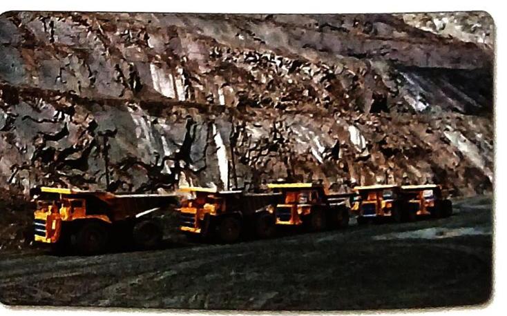 A line of large, yellow mining trucks sits in front of a textured, rocky cliff face in an open-pit mine.