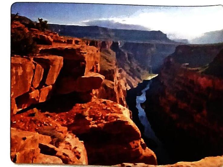 A wide canyon with steep, reddish-orange cliffs dominates the foreground. A blue river snakes through the deep canyon below, bordered by shadowed rock walls. Distant layered mountain ranges fade into a hazy sky where sunlight breaks through clouds.