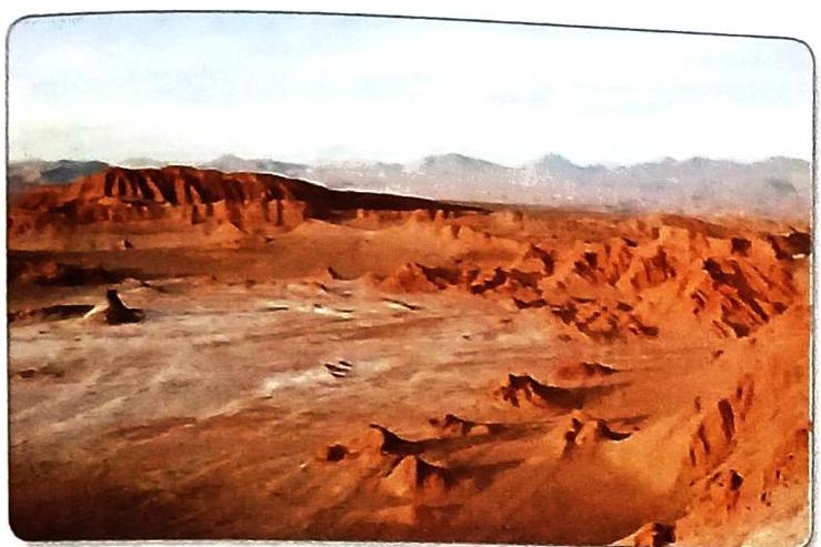 Rugged, eroded red rock formations characterize a desert landscape. A wide, sandy plain stretches towards these formations, with more rocky and uneven terrain in the foreground. Hazy mountains are visible on the horizon under a pale sky.