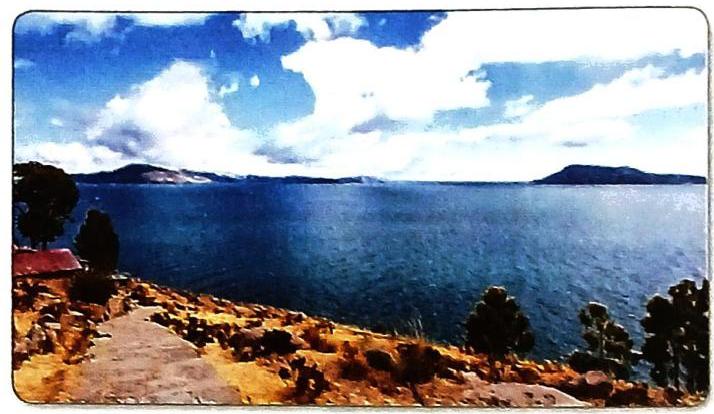 A wide blue body of water stretches to a horizon marked by distant, dark landforms. The sky above is bright blue, adorned with voluminous white clouds. In the foreground, a rugged, rocky slope with sparse vegetation and trees descends towards the water. A path winds through the landscape, and a small building with a red roof is situated on the left side.
